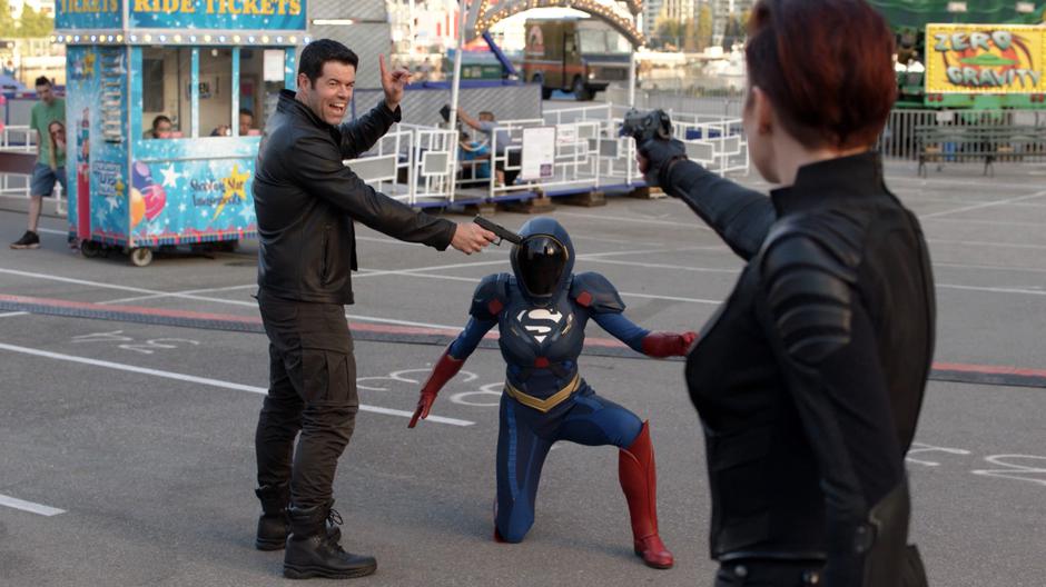 Otis Graves holds up his finger to stop Alex from shooting him while he holds his gun to Kara's suited head.