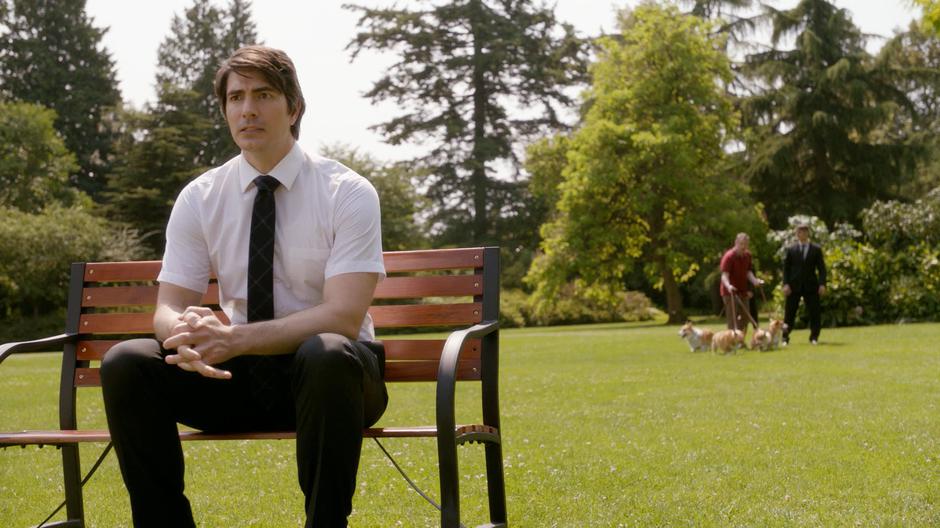 Ray sits on the bench in the middle of the park while the dog walker and his guards walk in the background.