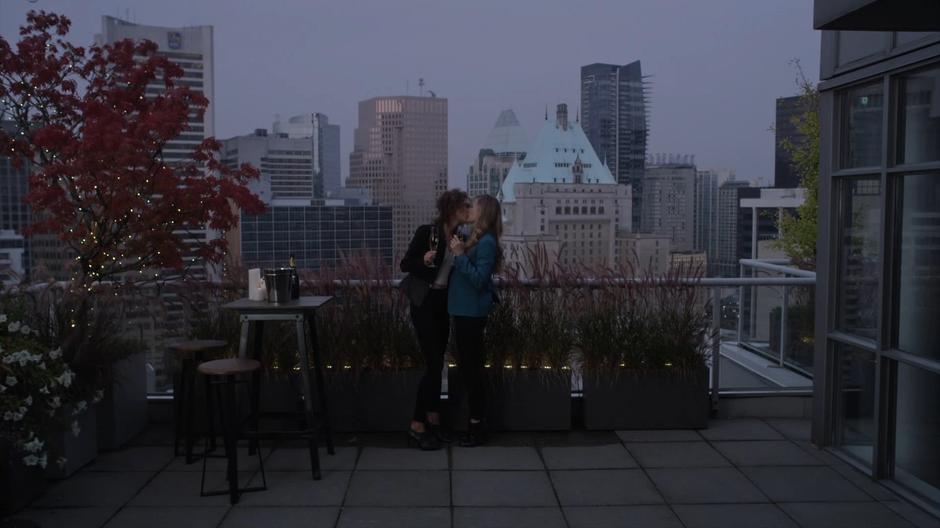 Kylie kisses Emma while they stand on the balcony at dusk.
