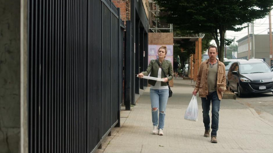 Izzy walks back to her apartment carrying a pizza next to Ben who is carrying the liquor.