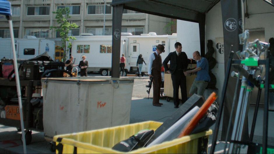 A props guy talk to two actors in the foreground while Trish and Jessica approach a trailer behind them.