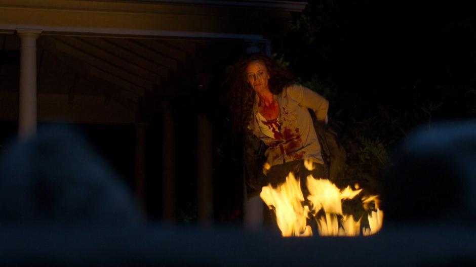 The mysterious woman takes off her jacket while covered in blood to throw it into the fire.