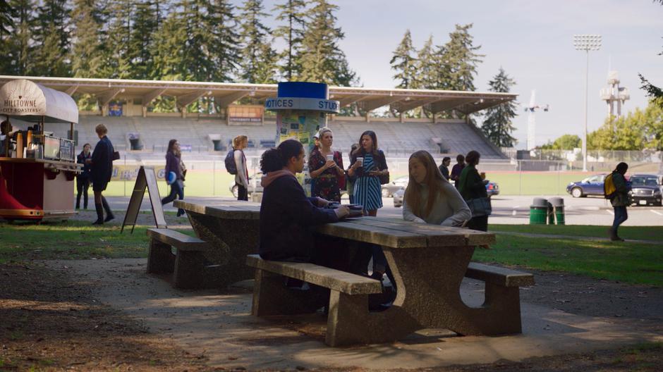 Maggie talks to Lucy as they walk away from the coffee stand with their drinks.