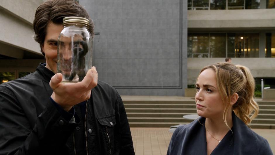 Sara looks over at Ray as he holds up a miniature Grodd in a jar.