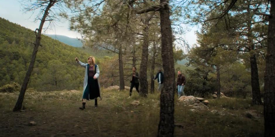 The Doctor leads Yasmin, Ryan, and Graham through the woods away from the TARDIS.