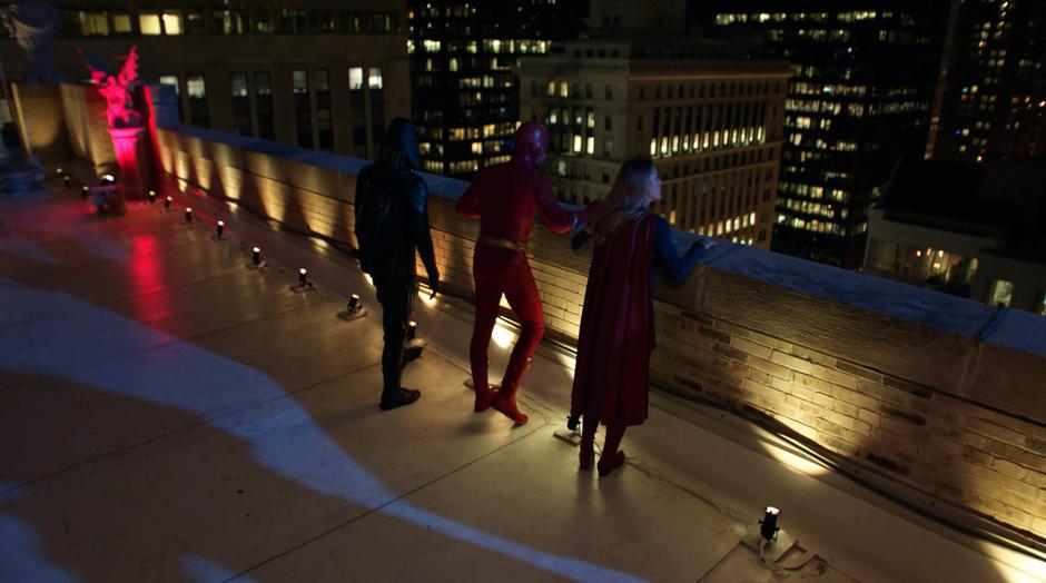Barry, Oliver, and Kara look over the edge of the roof to the streets of Gotham City.