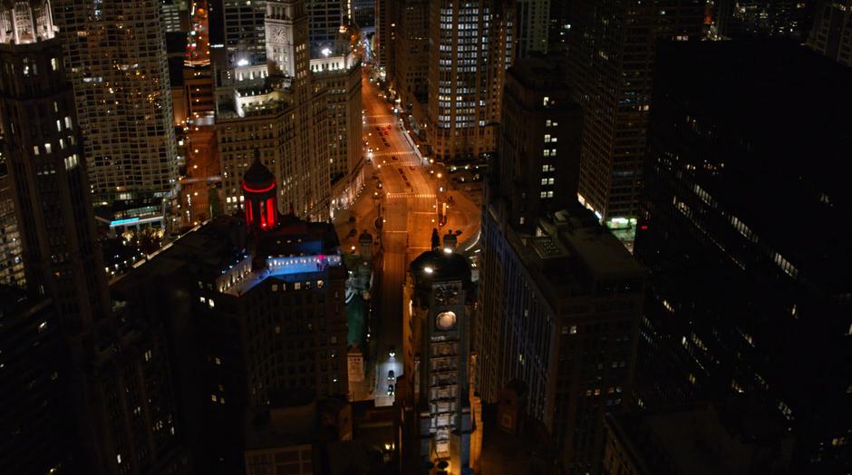 Batwoman looks down from the Wayne Enterprises tower at the rooftop where Kara, Oliver, and Barry just uncovered the Bat-Signal.