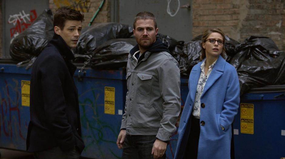 Barry, Oliver, and Kara turn around and watch as a rich couple is escorted by heavily armed guards through the alley.