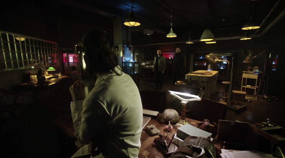 Dr. Deegan sits on his desk as Oliver and Diggle enter to talk to him.