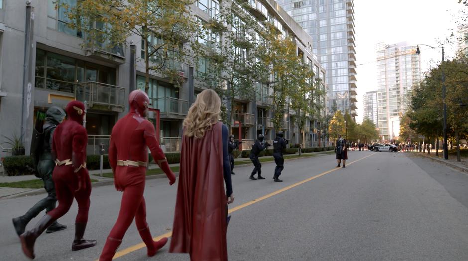 Barry, Earth-90 Flash, Oliver, and Kara walk down the street toward the Monitor while the police surround him.
