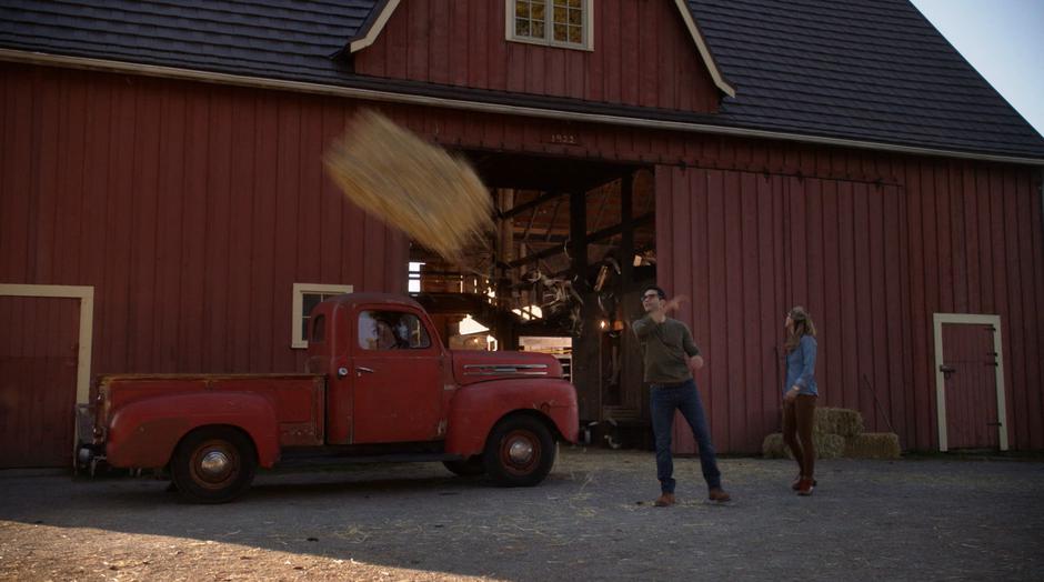 Clark throws a bail of hay while talking to Kara.