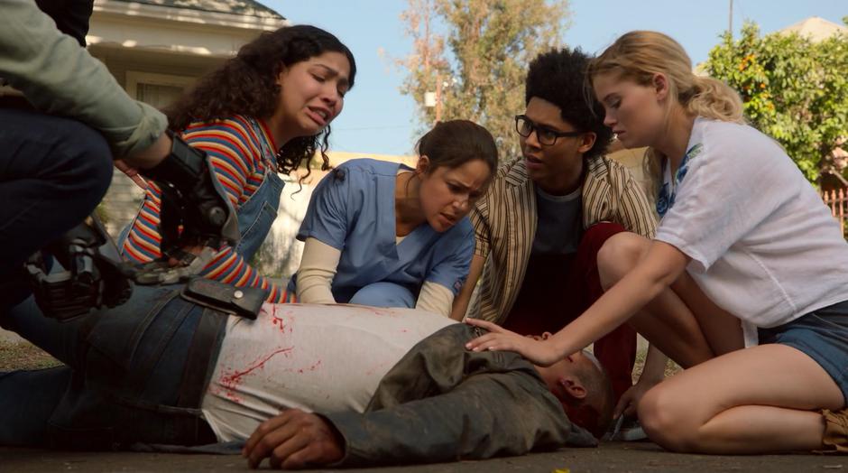 Molly, Sofia, Alex, and Karolina kneel over Topher as he lies bleeding in the street.