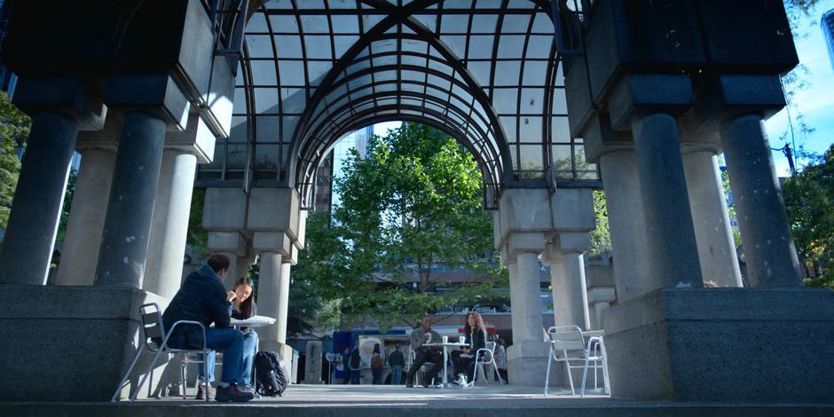 Jeff and Carly laugh while sitting at a table with coffee under a coversed structure.