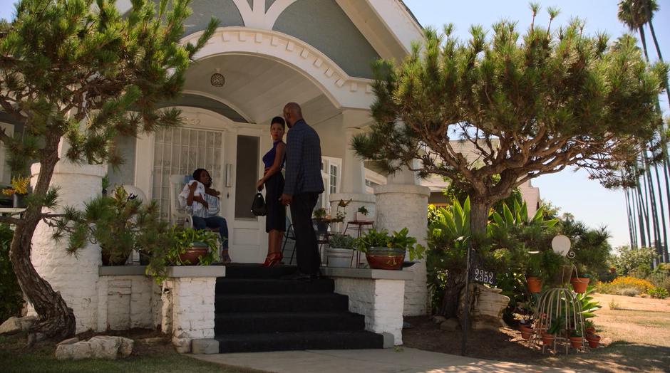 Catherine glances back at Geoffrey while they stand on the porch talking to Tamar who is holding her baby.