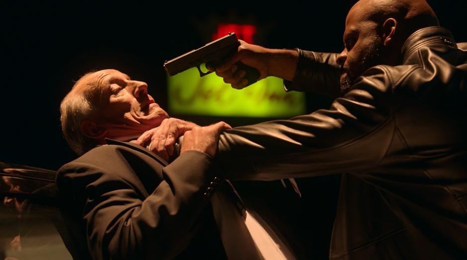 Geoffrey holds Detective Flores by the throat while pointing his own gun at his head.