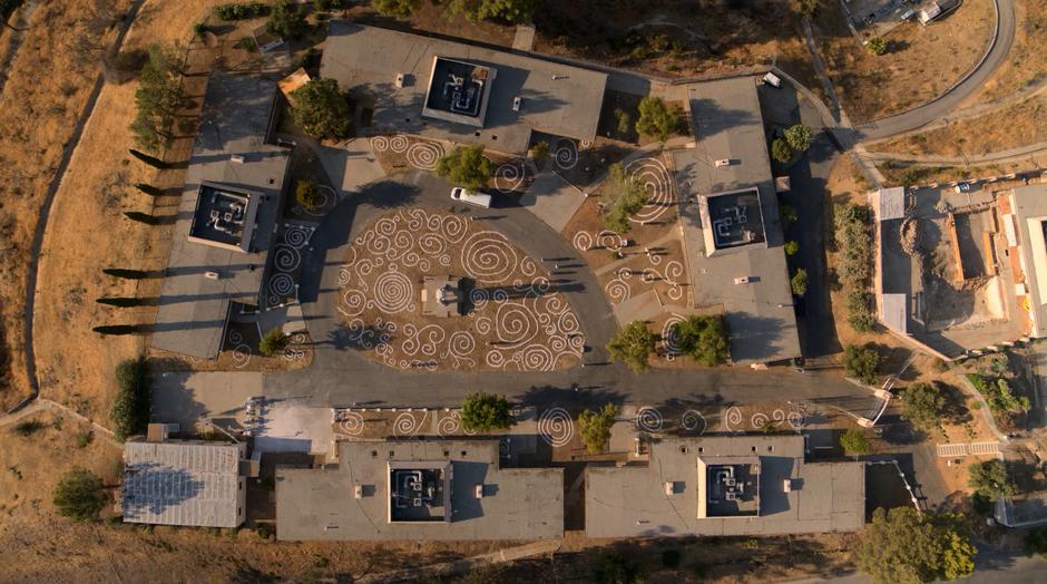 An overhead establishing view of the compound with spiraling symbols on the ground made of white rocks.
