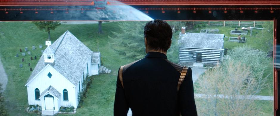 Pike stands in front of the bridge screen looking at an image of the village church from space.