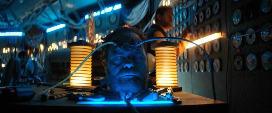 A disembodied head of Harry Mudd lies on a table with wires running out of it while other duplicates of him work in the background.