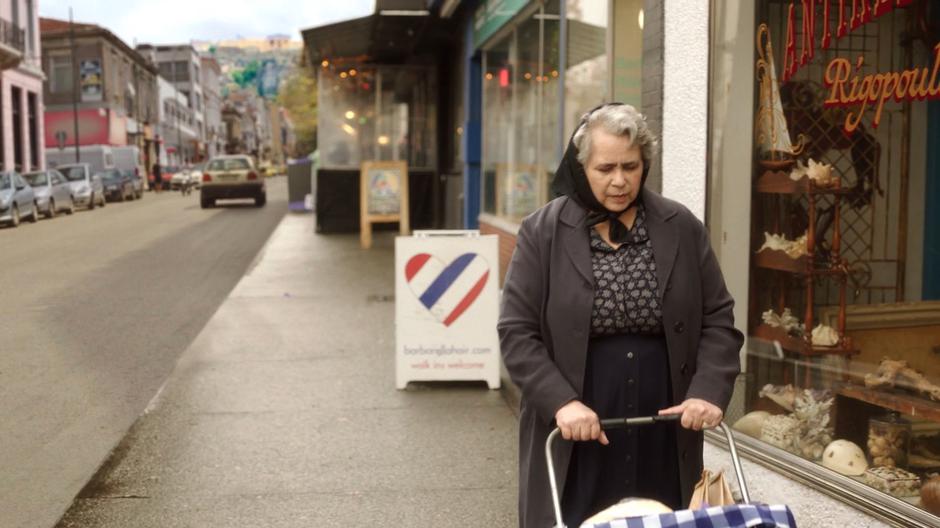 An old woman walks down the street past the front of the store.