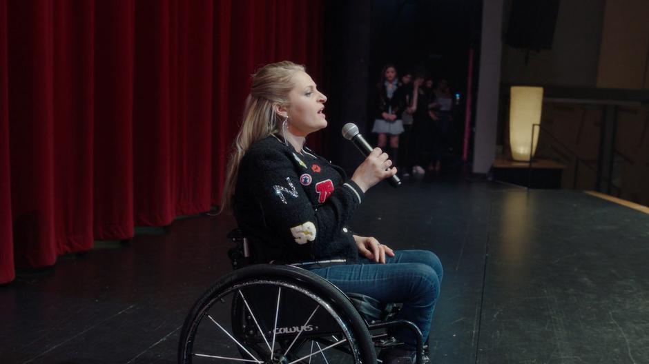 Maggie watches from the side of the stage as one of the prospective singers auditions on stage in a wheelchair.