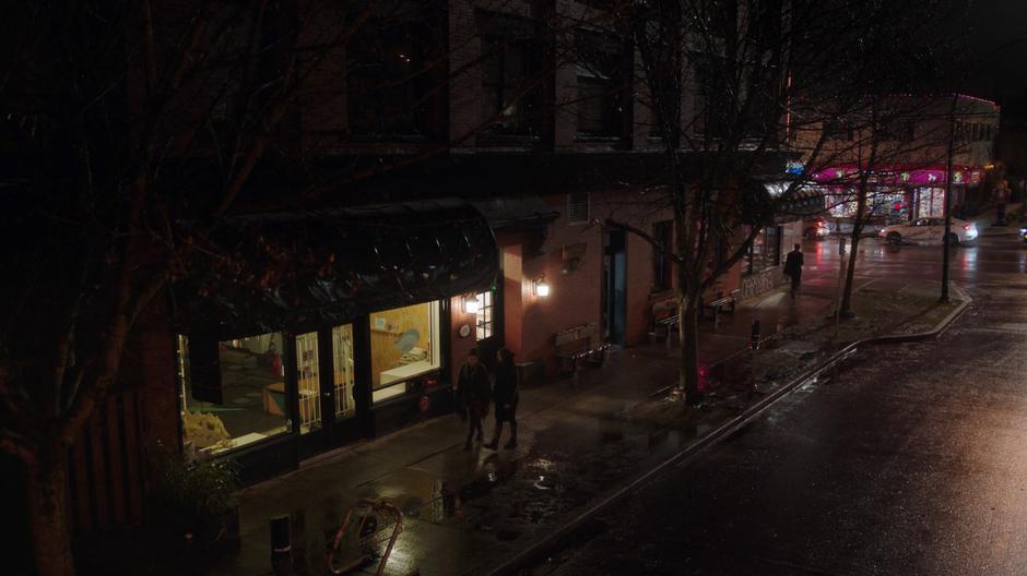 Mel and Jada walk down the sidewalk at night in the rain.