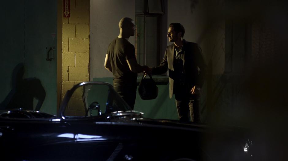 Lucifer exchanges a duffel bag with a guy behind a building at night.