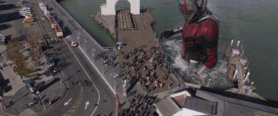 A gigantic Ant-Man walks up to the pier in the water.