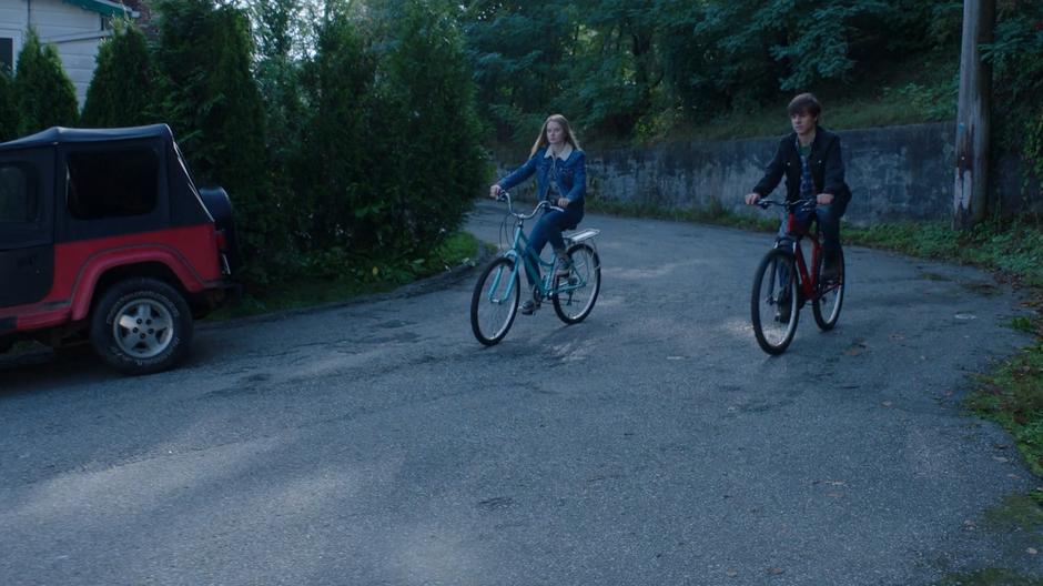 Two kids ride their bikes down the street.