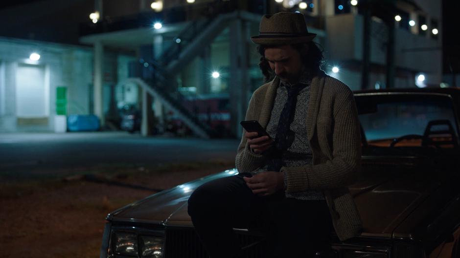 Noah looks at the stills from the video on his phone while leaning against his car.