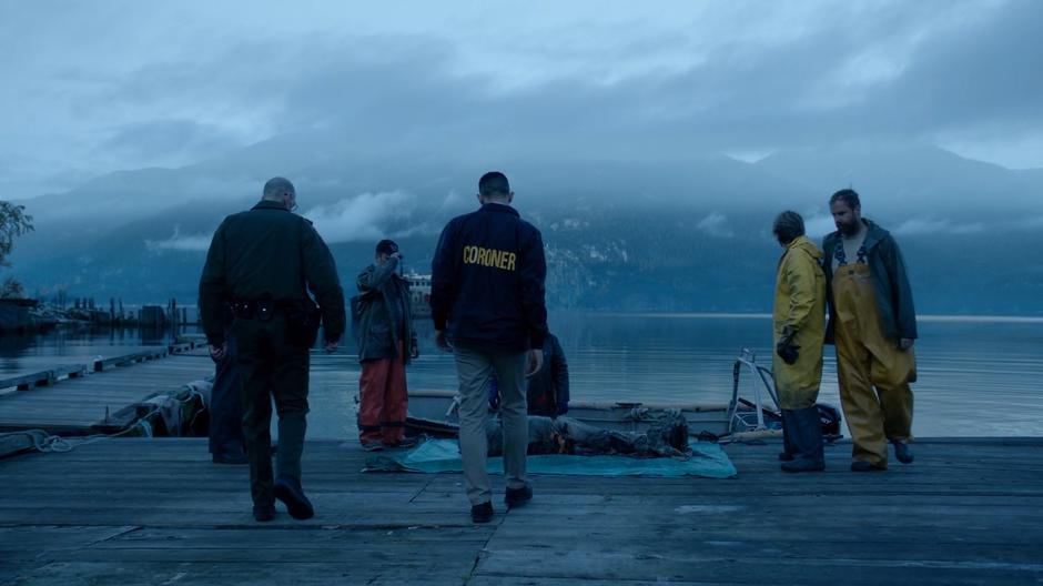 The coroner walks over the body which is surrounded by serveral fishermen.