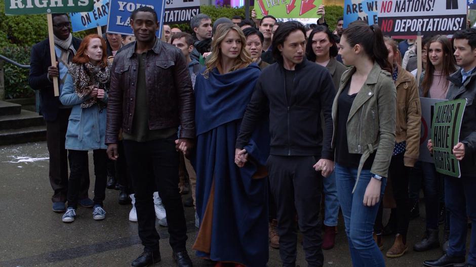 J'onn, Kara, Brainy, and Nia join hands at the front of the protest.