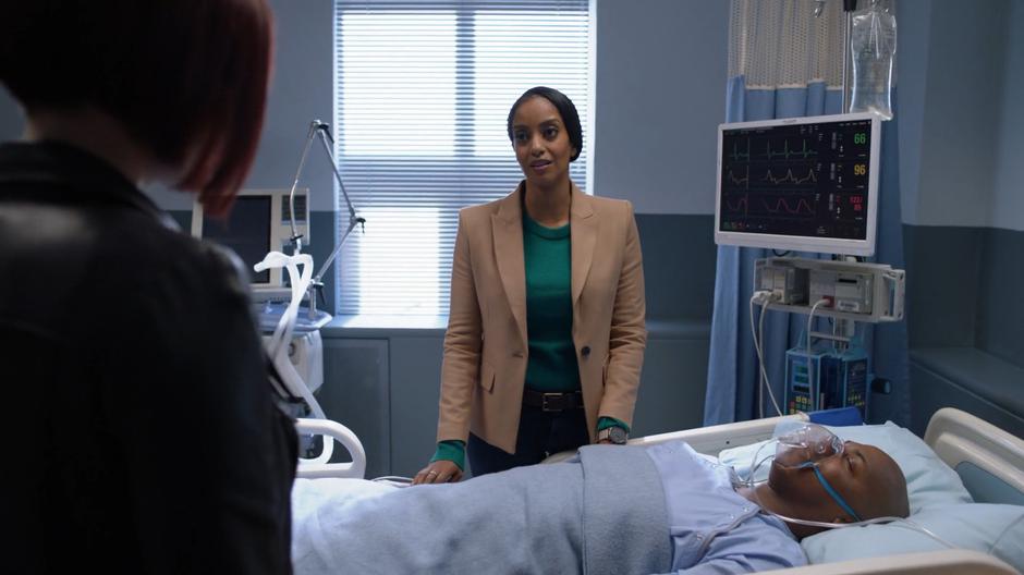 Kelly talks to Alex while they stand over James's hospital bed.