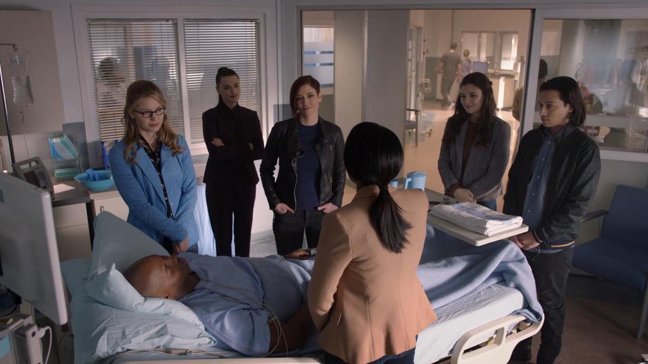Kara, Lena, Alex, Nia, and Brainy stand around James's hospital bed with Kelly.