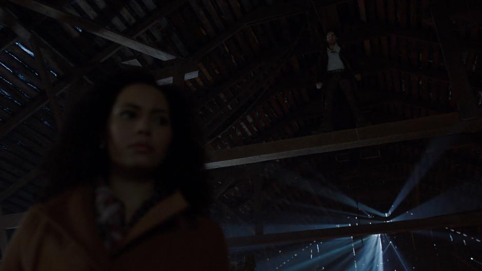 Macy looks around the loft while Viralis stands on a roof beam above her.