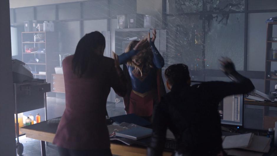 Lena, Kara, and Alex duck as the glass wall explodes behind them.