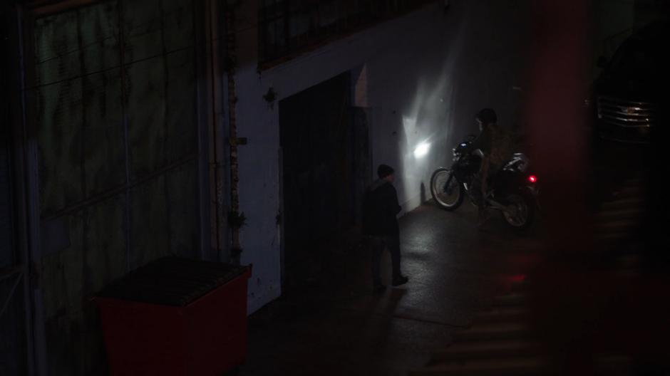 The courier parks his motorcycle in front of the warehouse while the guard walks over to him.