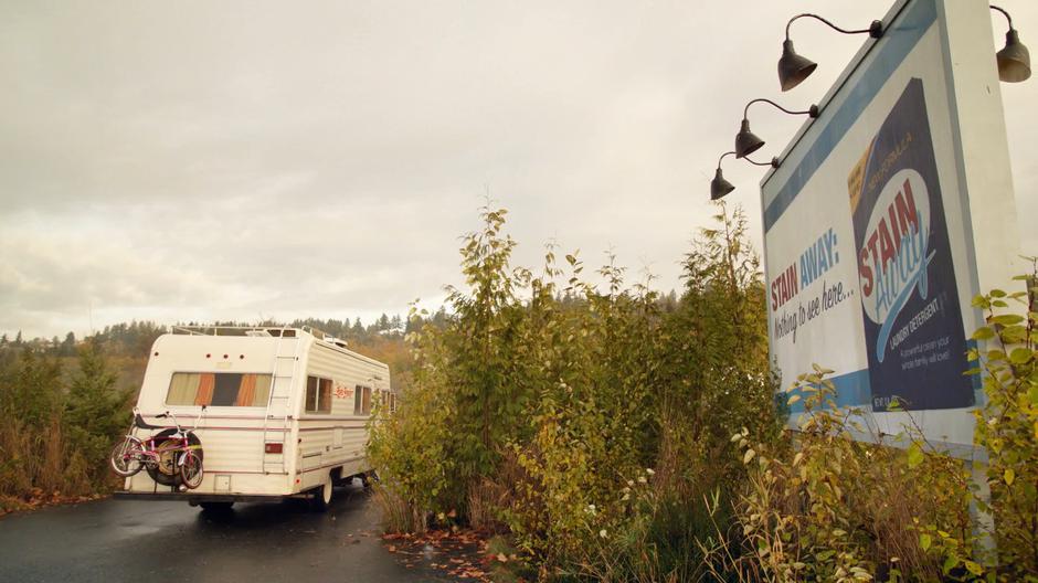 The RV pulls to a stop behind some plants next to a billboard.