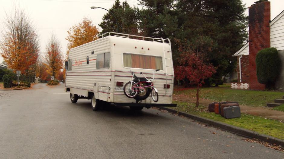 The RV drives off leaving a line of suitcases on the sidewalk.