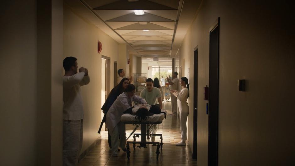 Margo walks with Professor Lipson and a nurse as they push a gravely injured Eliot down the hallway.