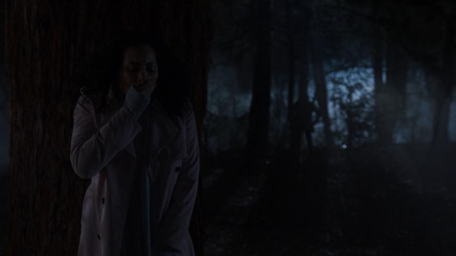 Macy covers her mouth while hiding behind a tree to avoid detection from the demon-controlled Galvin searching the woods behind her.