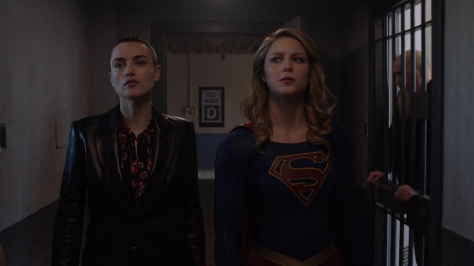 Lena and Kara walk down the D Block hallway past cells full of inmates.