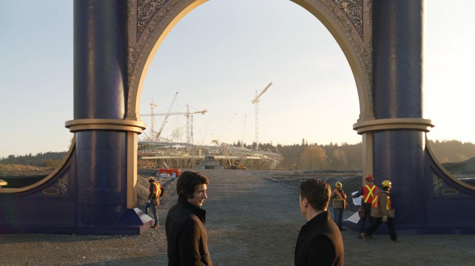 Ray looks over at Nate as they stand in front of the theme park gate while a large enclosure is constructed in the distance.