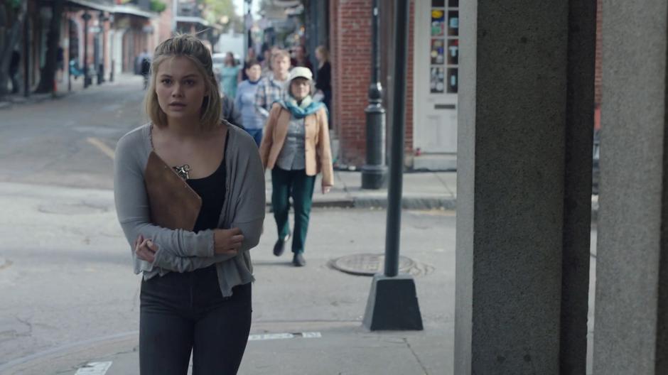 Tandy hugs herself while walking down the sidewalk with a clipboard looking for a mark.