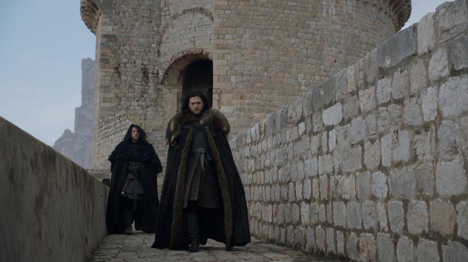 Jon walks down the wall escorted by two guards.