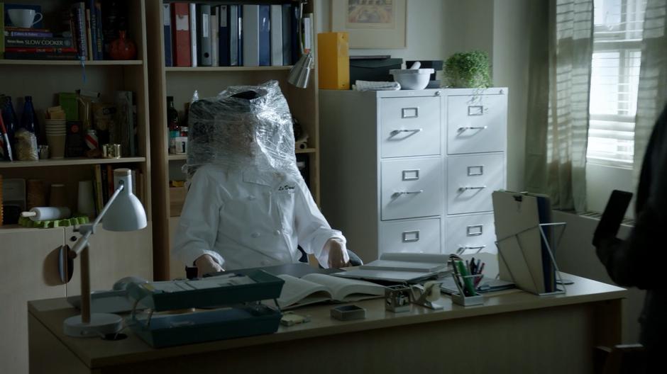 Chef's body sits behind her desk with her head covered in plastic wrap.