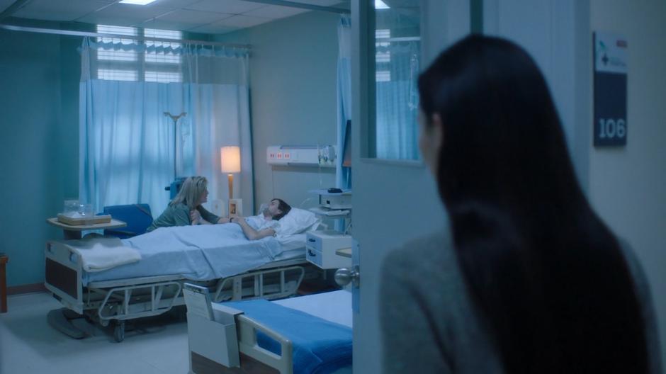 Ryn glances into the room where a woman is leaning over a patient.