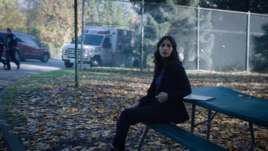 Helen sits at a picnic table outside and turns as Ben & Maddie approach.