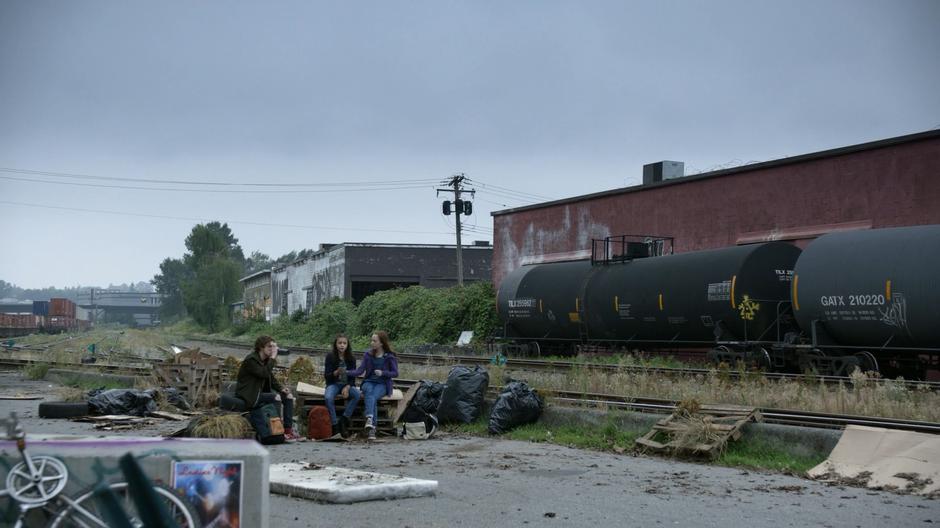 Oliver, Pippi, and Annie sit on pallets in the alley drinking water and waiting for Baron.