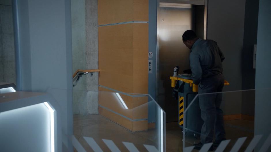 Clive waits at the elevator disguised as a janitor.