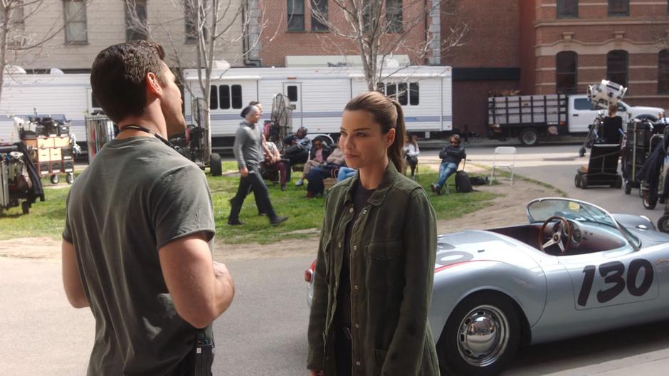 Chloe talks to a crew member in front of Lucifer's car which is parked at circus.